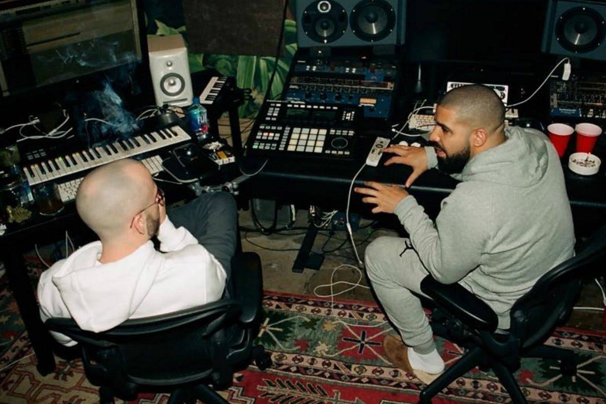 Two music producers working together in a recording studio, surrounded by keyboards, speakers, and mixing equipment, discussing a project at a workstation.