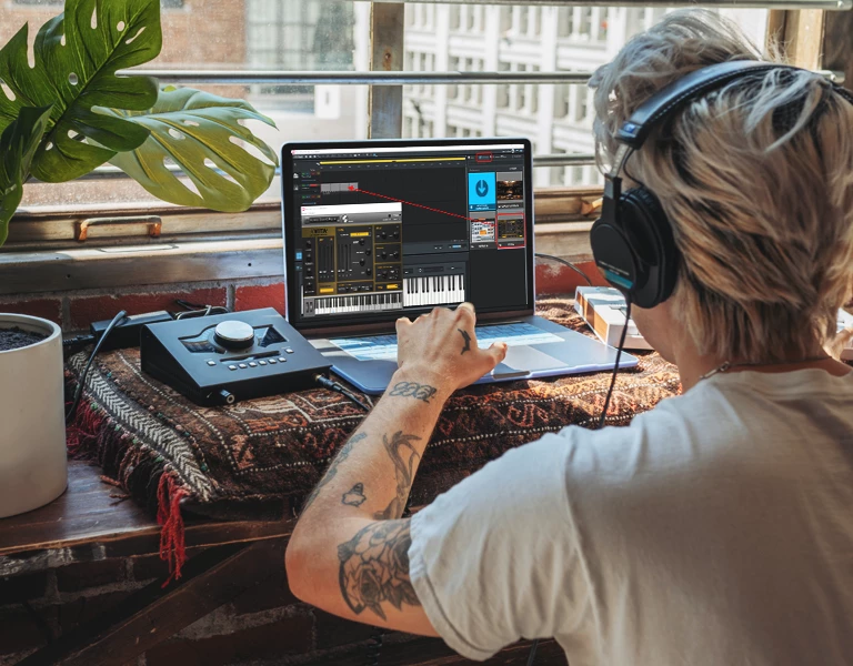 Person with tattoos and headphones looking at a laptop and small audio interface, making a beat.