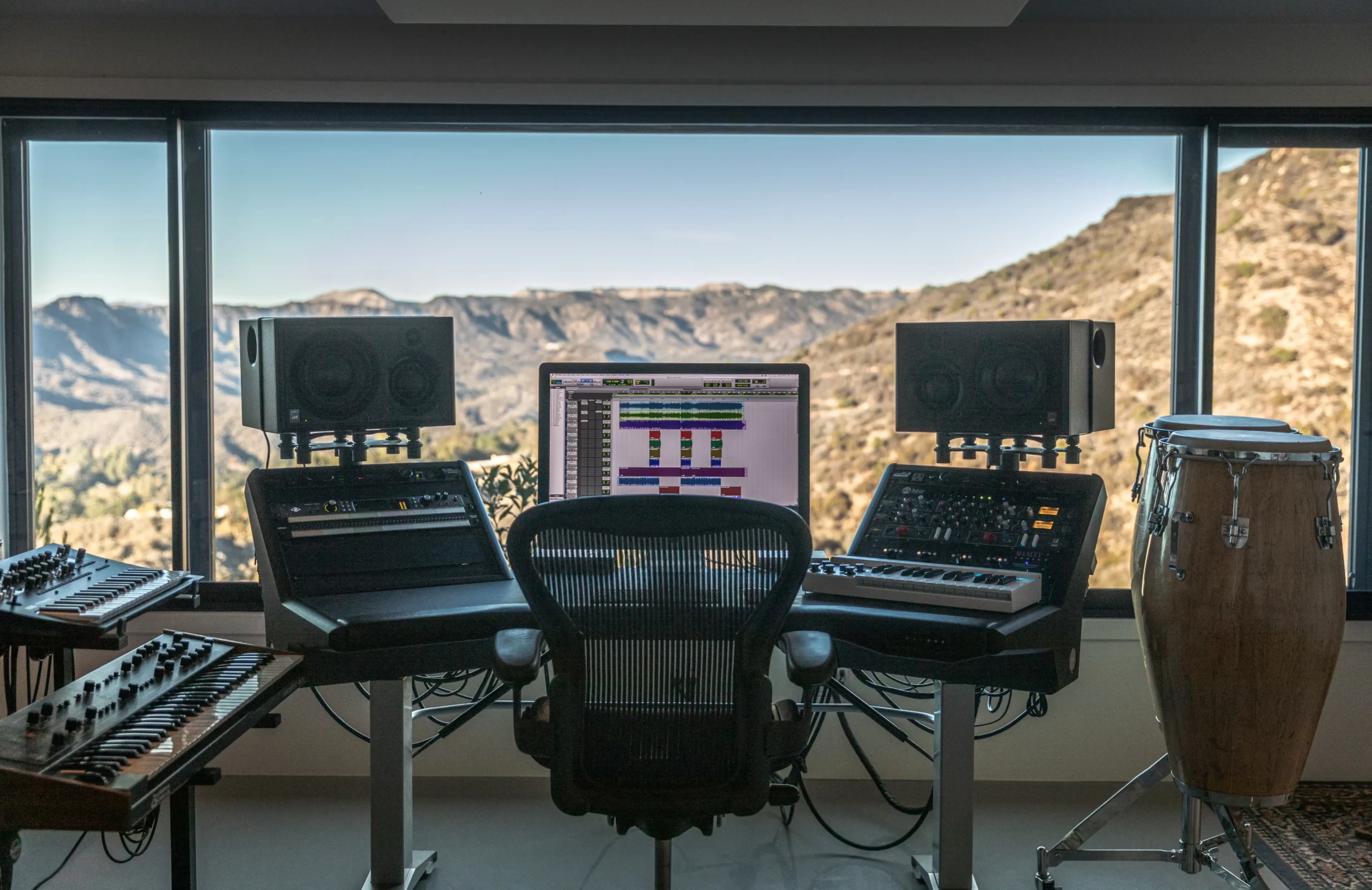 A professional home recording studio desk with a computer monitor, keyboard, and speakers overlooking a scenic mountain view.